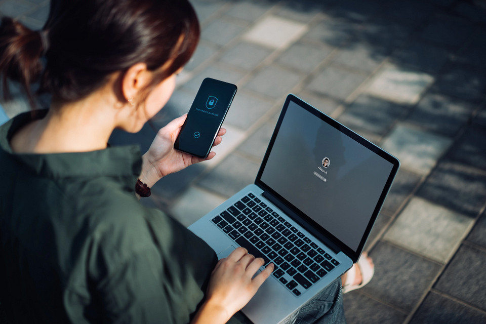 Jonge Aziatische zakenvrouw zit op een bankje in een stadspark buiten te werken, logt in op haar laptop en houdt een smartphone in haar hand met een pictogram van een veiligheidssleutelslot op het scherm. Privacybescherming, internet en mobiel beveiligingsconcept