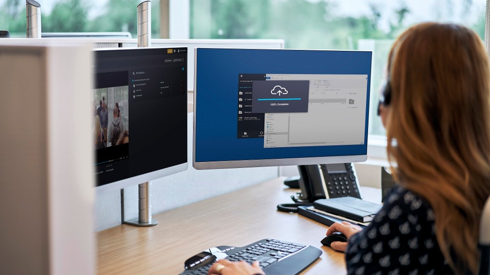 An operator at a desk uses dual monitors with cloud and video applications, running a flexible hybrid cloud security deployment.