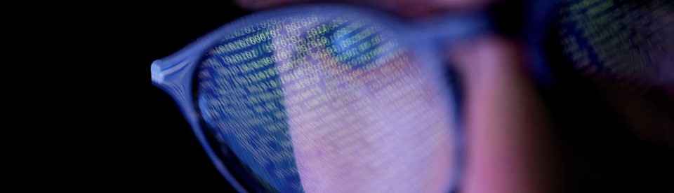 Een persoon met een bril in een donkere kamer kijkt naar een computerscherm waarop codes of programmeerwerk worden getoond, met reflecties op de bril