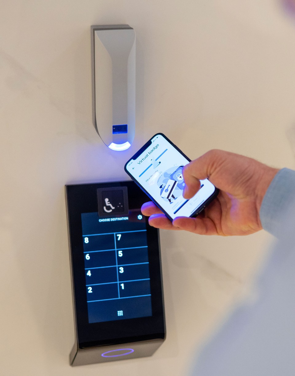 Una mano sosteniendo un dispositivo móvil cerca de un panel de control de acceso con pantalla táctil. La pantalla muestra una credencial móvil y se está utilizando para desbloquear el acceso al edificio.