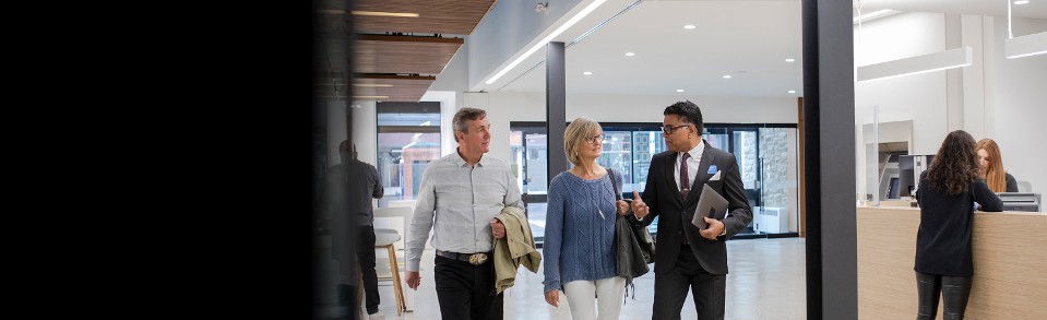 Ein Bankmanager führt zwei Kunden durch eine Banklobby und zeigt dabei moderne Banksicherheit.